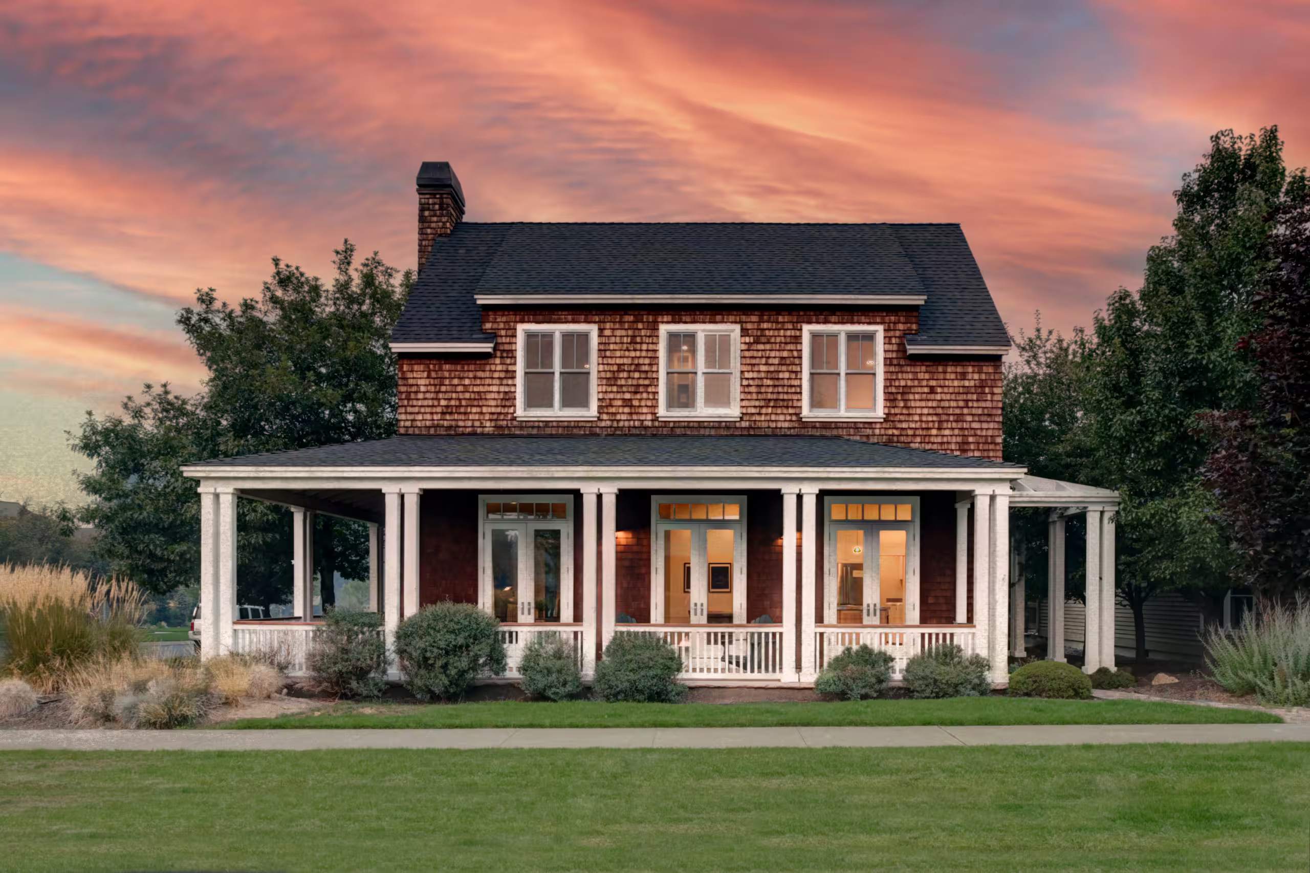 Cedar-shingle farmhouse with a white wraparound porch across a wide green lawn at dusk