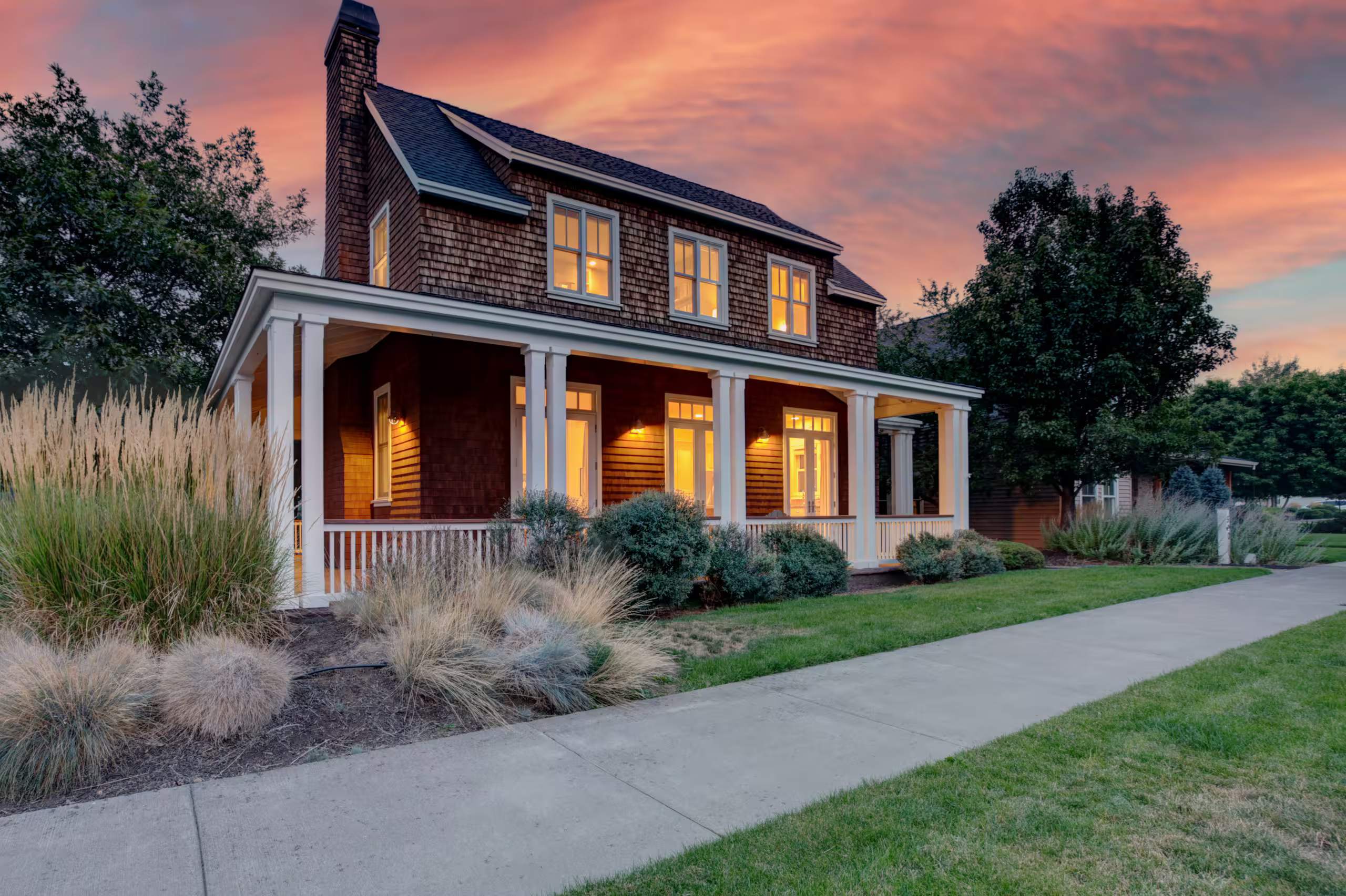 Cedar-shingle farmhouse with a white-columned wraparound porch glowing at dusk